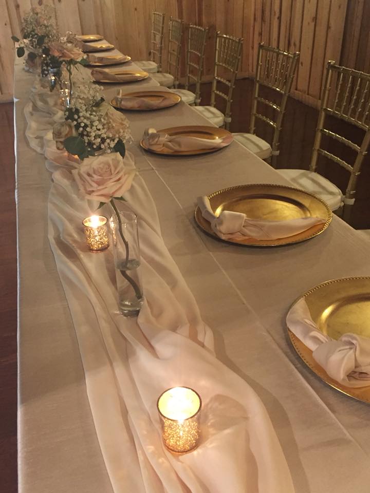table scape with a folded napkin laying on top of the gold charger plate. Candles and white roses adorn the center pieces